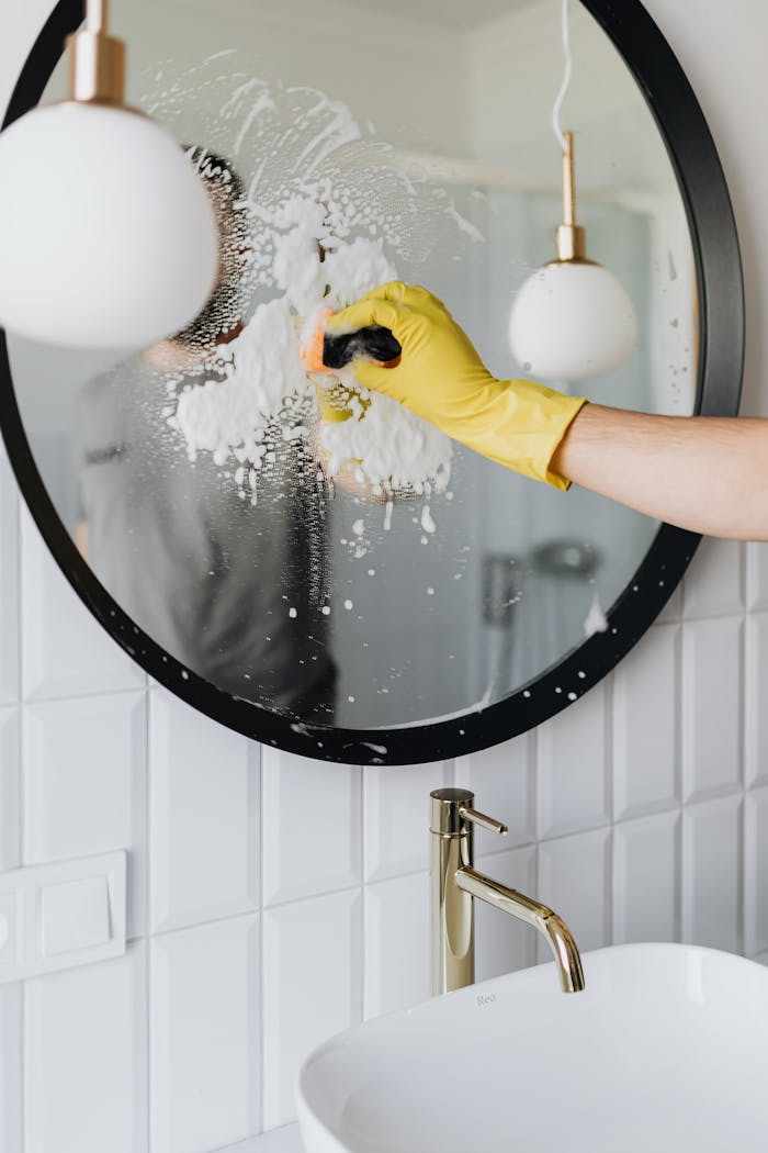 about-02 A person wearing a yellow glove cleans a bathroom mirror with foam spray.