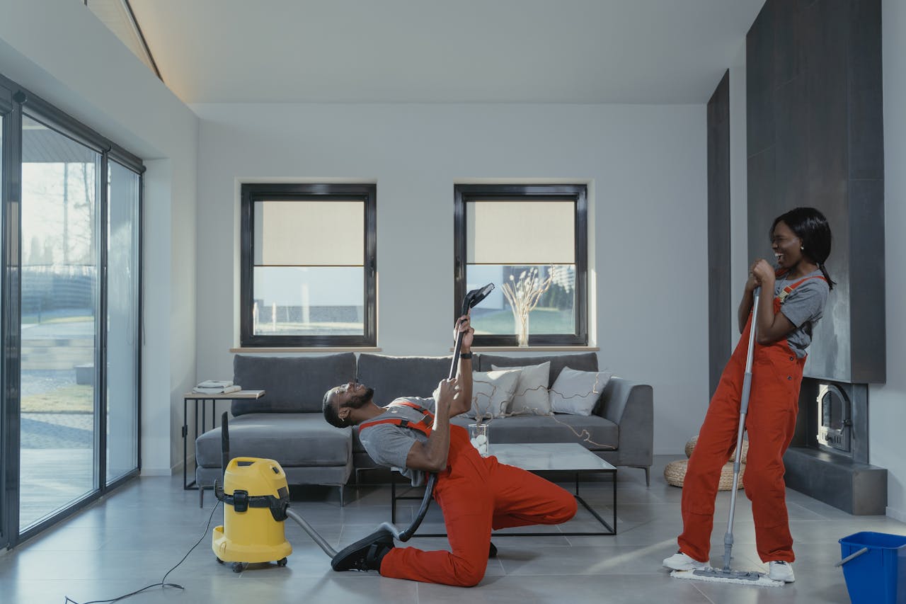 hero-01 Fun and engaging cleaning session with a man and woman in overalls using cleaning tools in a modern living room.